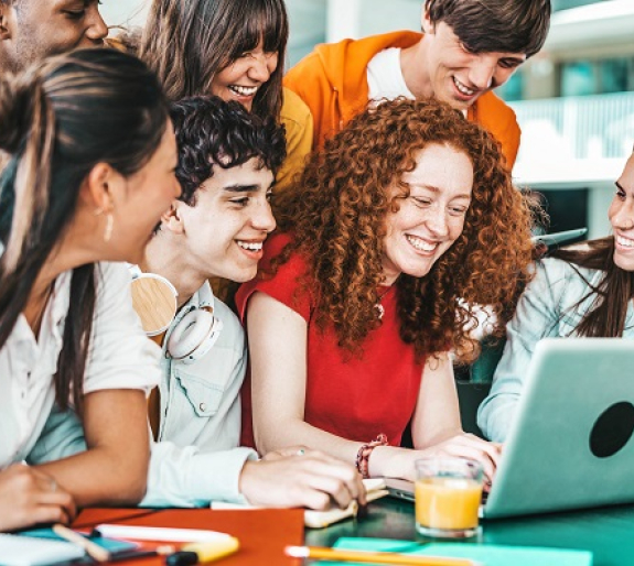 The image shows a diverse group of young people gathered closely around a laptop, smiling and engaging enthusiastically